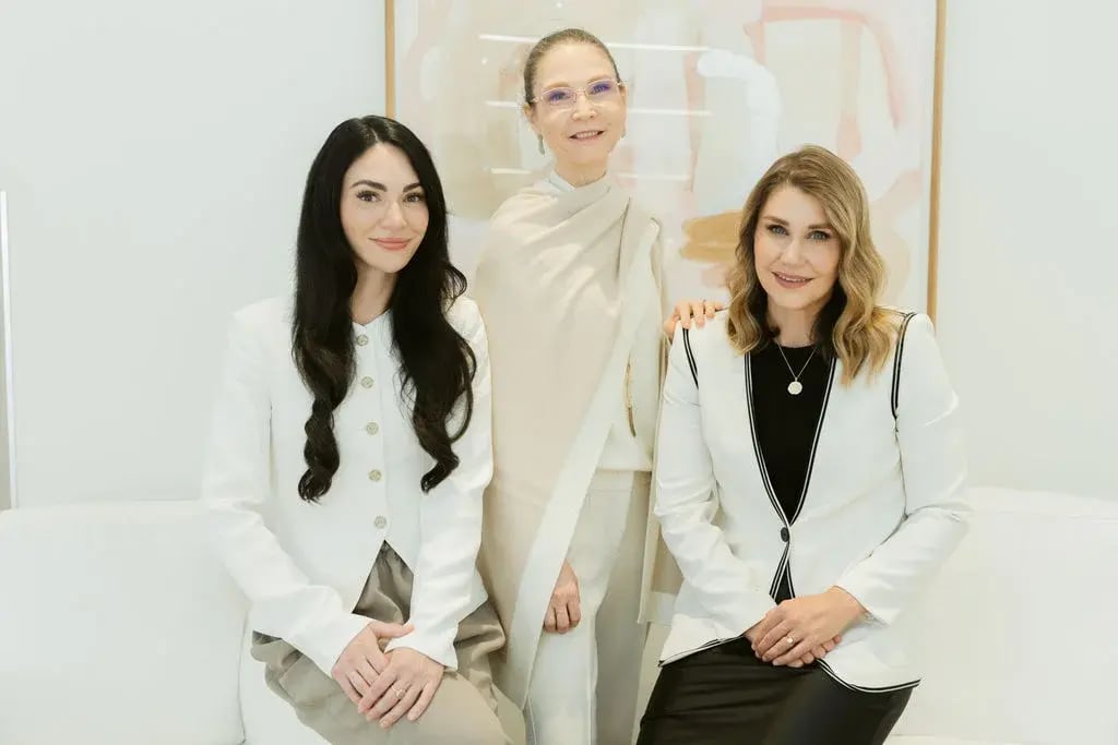 three-women-sitting-together-on-white-couch-wearing-light-colored-outfits-in-modern-office-with-abstract-art-in-background