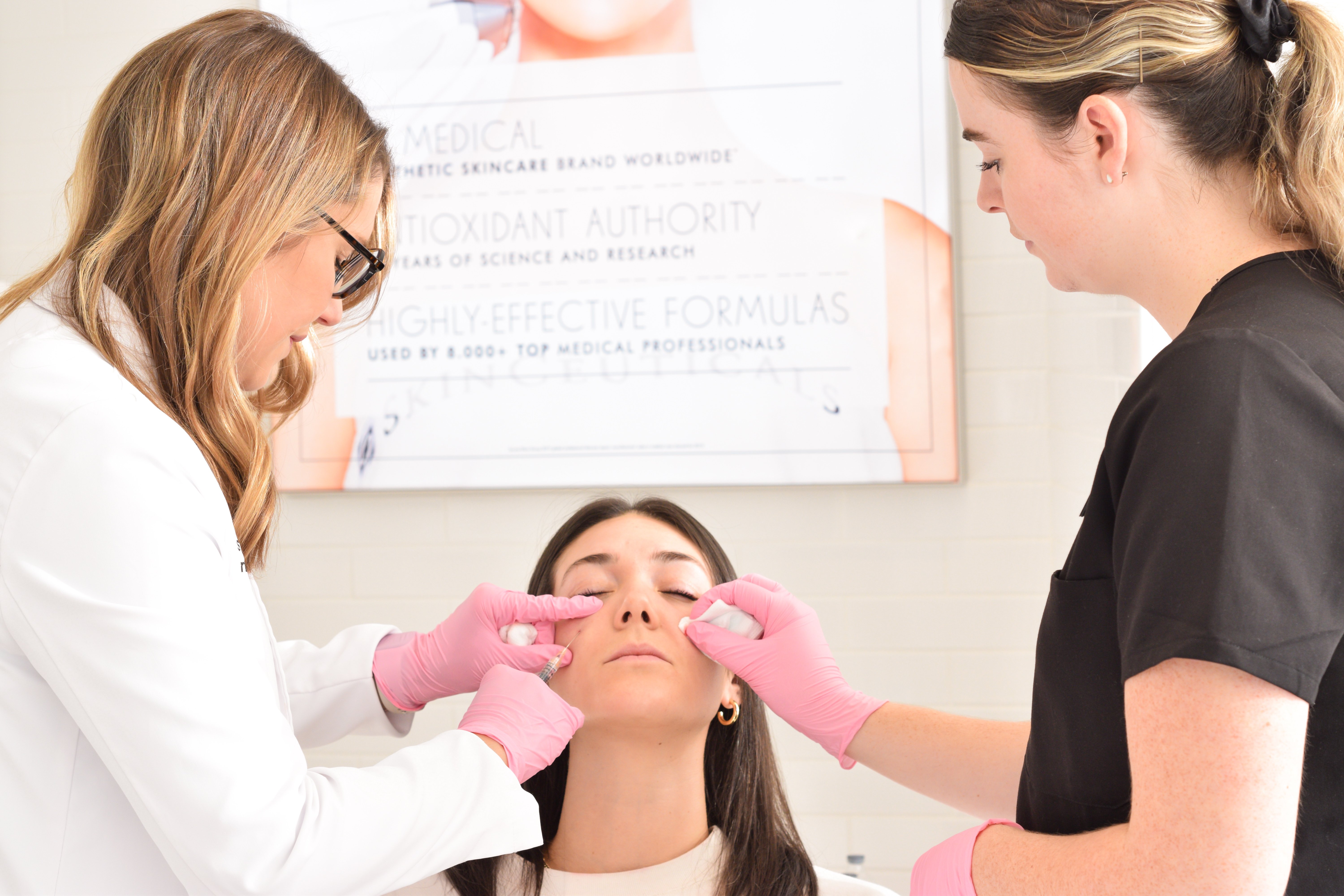 two-medical-professionals-in-white-and-black-uniforms-performing-dermal-fillers-victoria-bc-treatment-on-woman-sitting-with-eyes-closed-and-wearing-pink-gloves