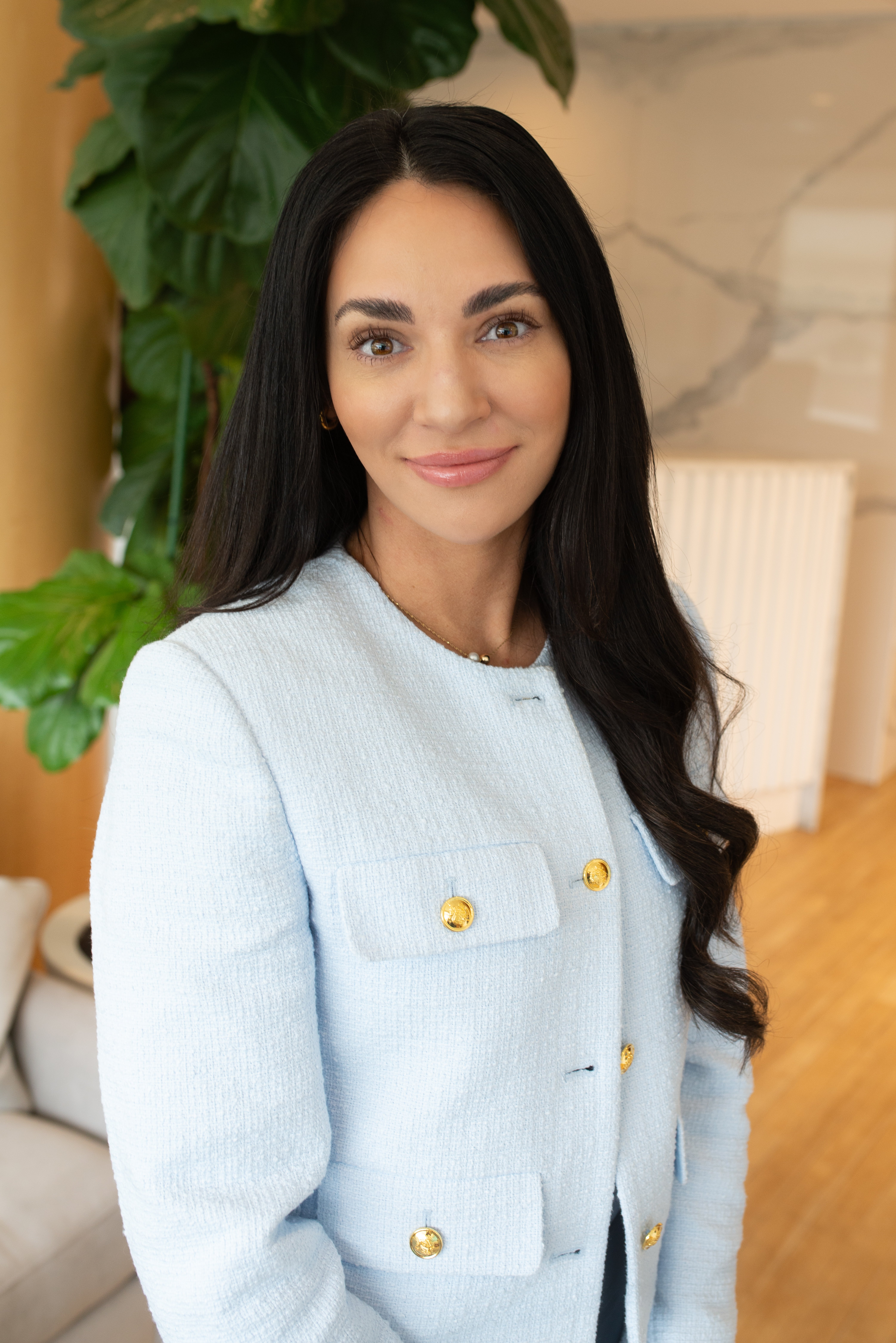 woman-with-long-dark-hair-wearing-light-blue-jacket-with-gold-buttons-standing-in-modern-office-with-green-plant-in-background