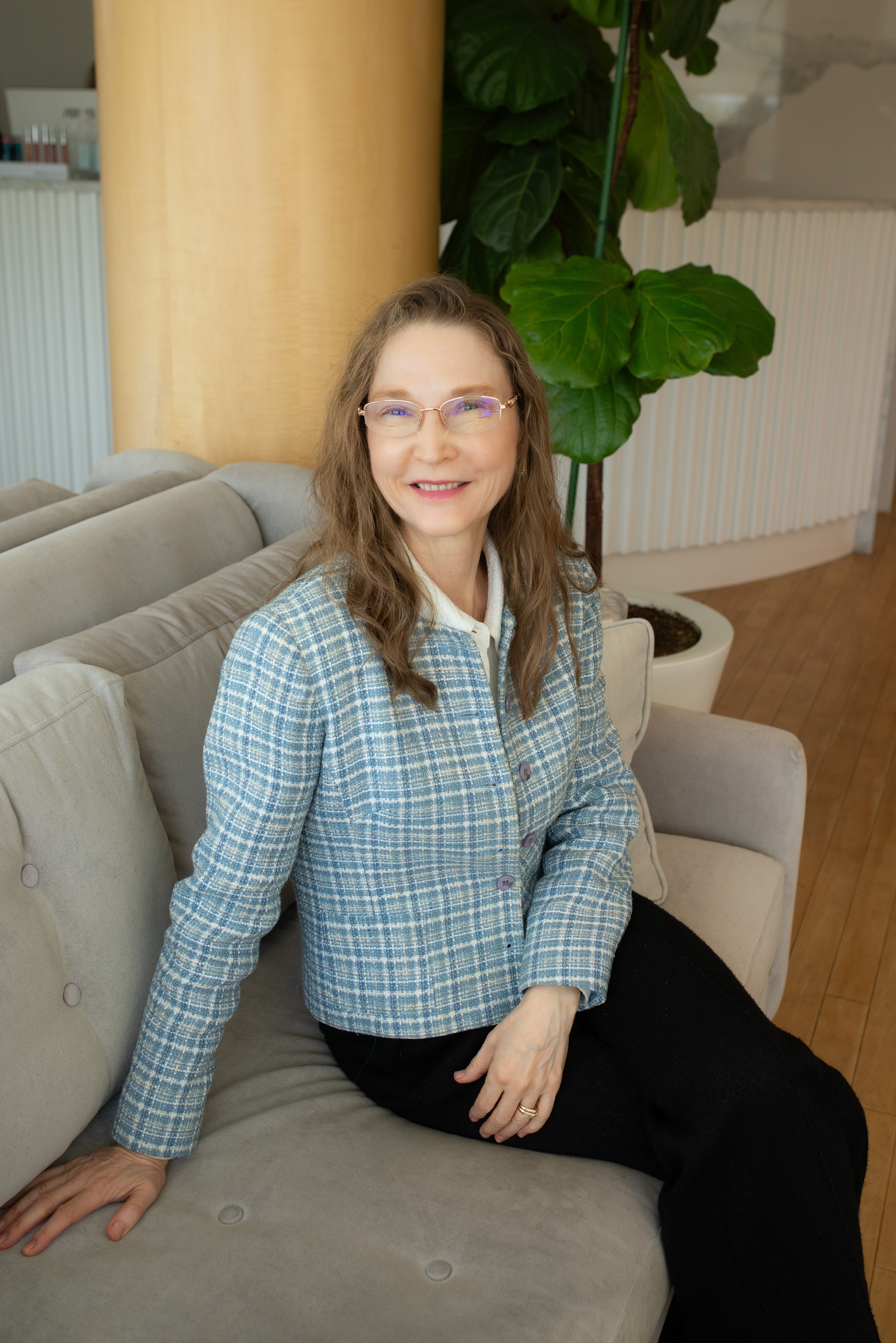 woman-with-long-light-brown-hair-wearing-blue-plaid-jacket-and-glasses-sitting-on-light-gray-couch-in-modern-office-with-green-plant-behind
