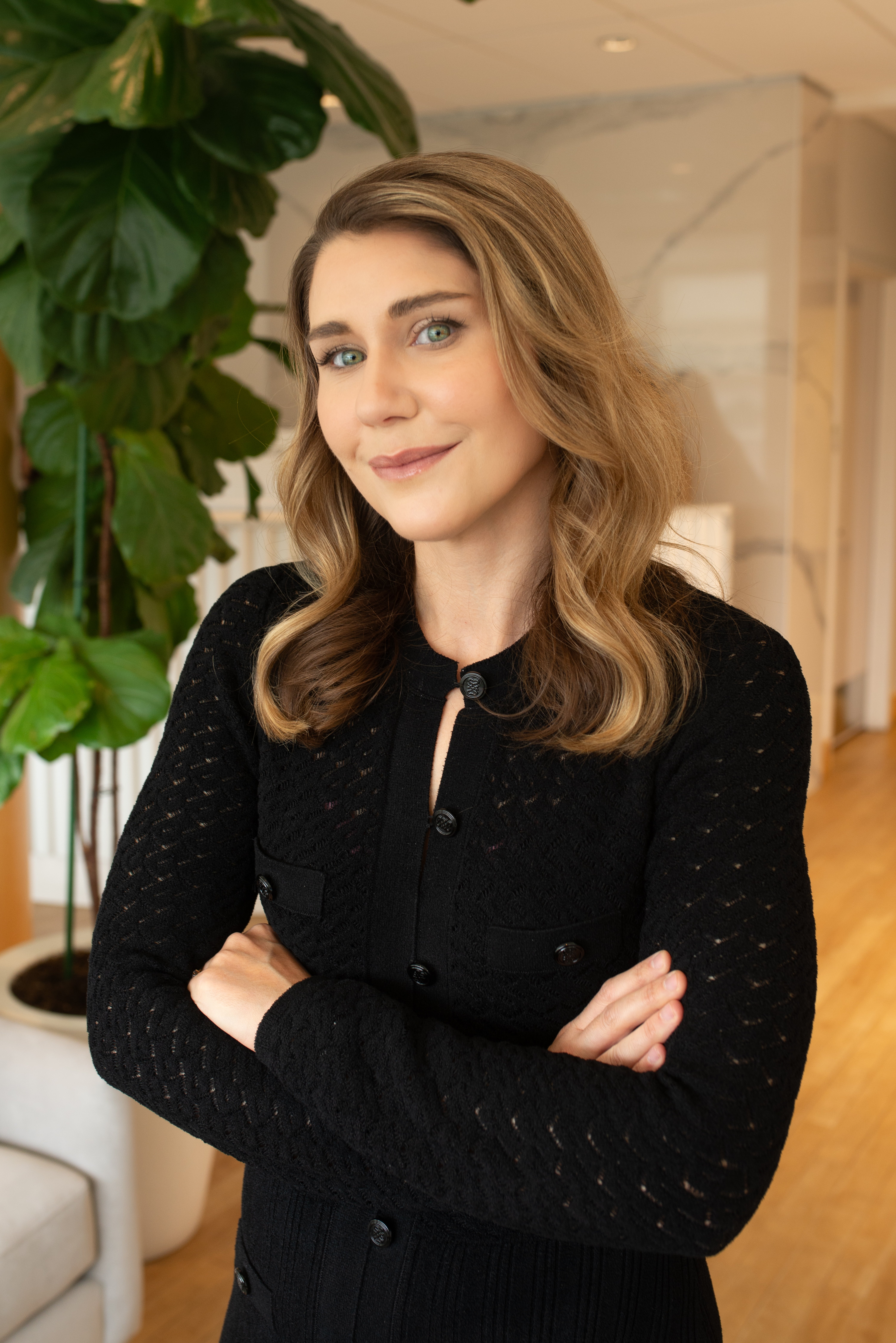 woman-with-wavy-blonde-hair-wearing-black-dress-standing-with-arms-crossed-in-modern-office-with-green-plant-in-background